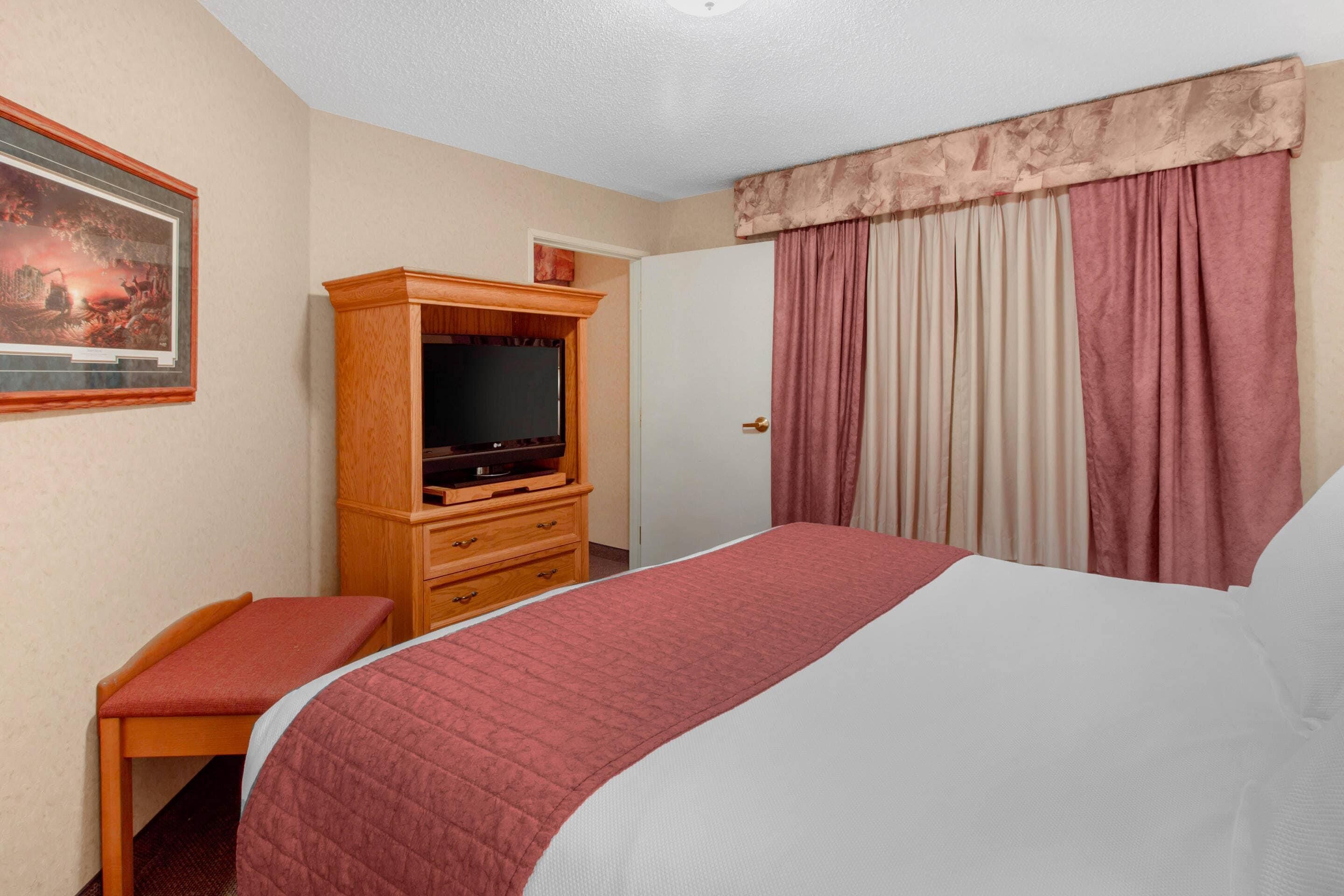A hotel room with a bed, a television on a wooden cabinet, a bench, and a painting on the wall.