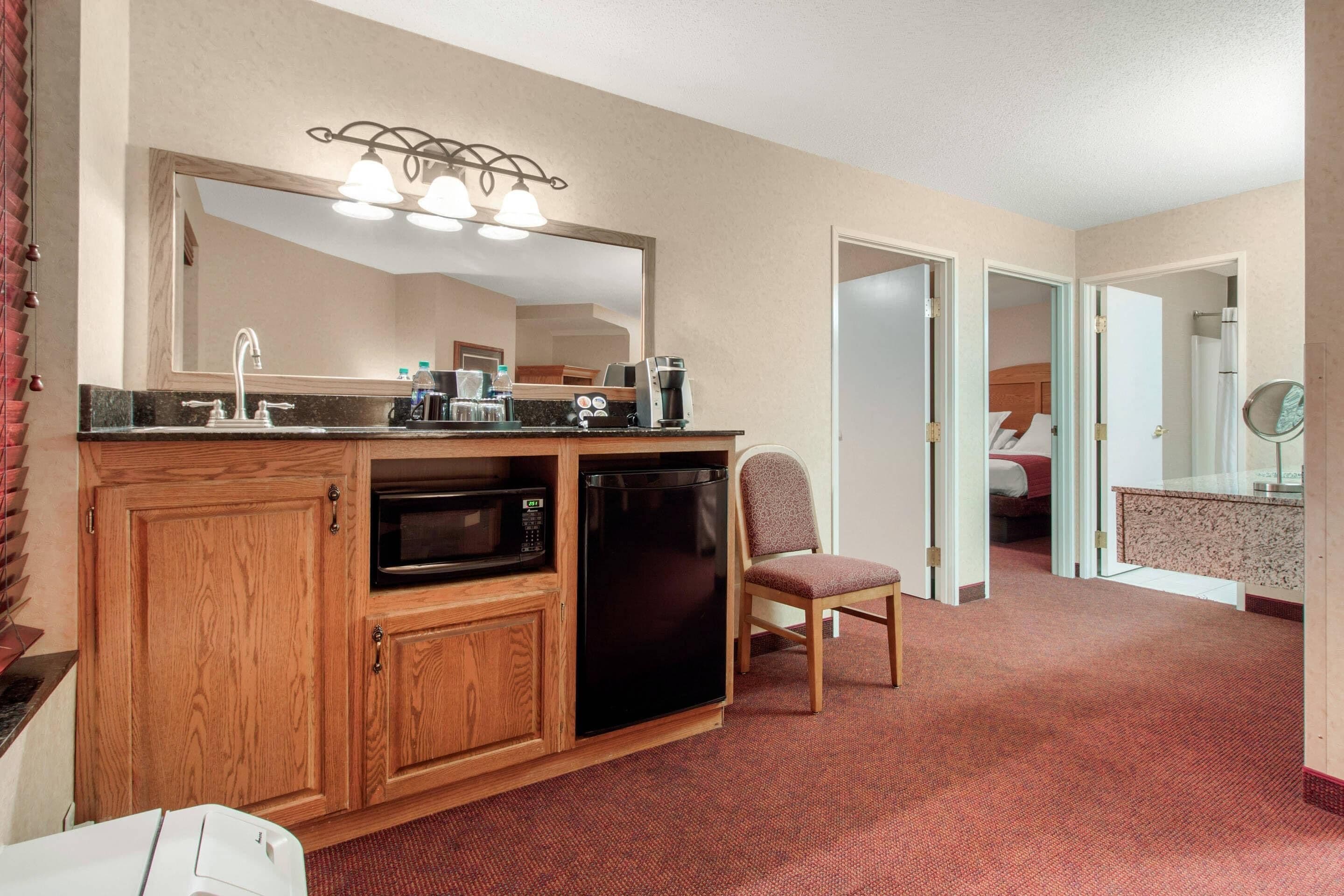 A compact hotel room with a kitchenette, a large mirror, a single chair, and a bed in the adjacent room.
