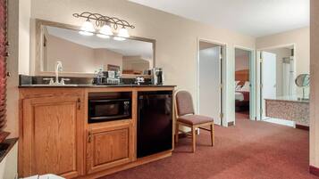 A compact hotel room with a kitchenette, a large mirror, a single chair, and a bed in the adjacent room.