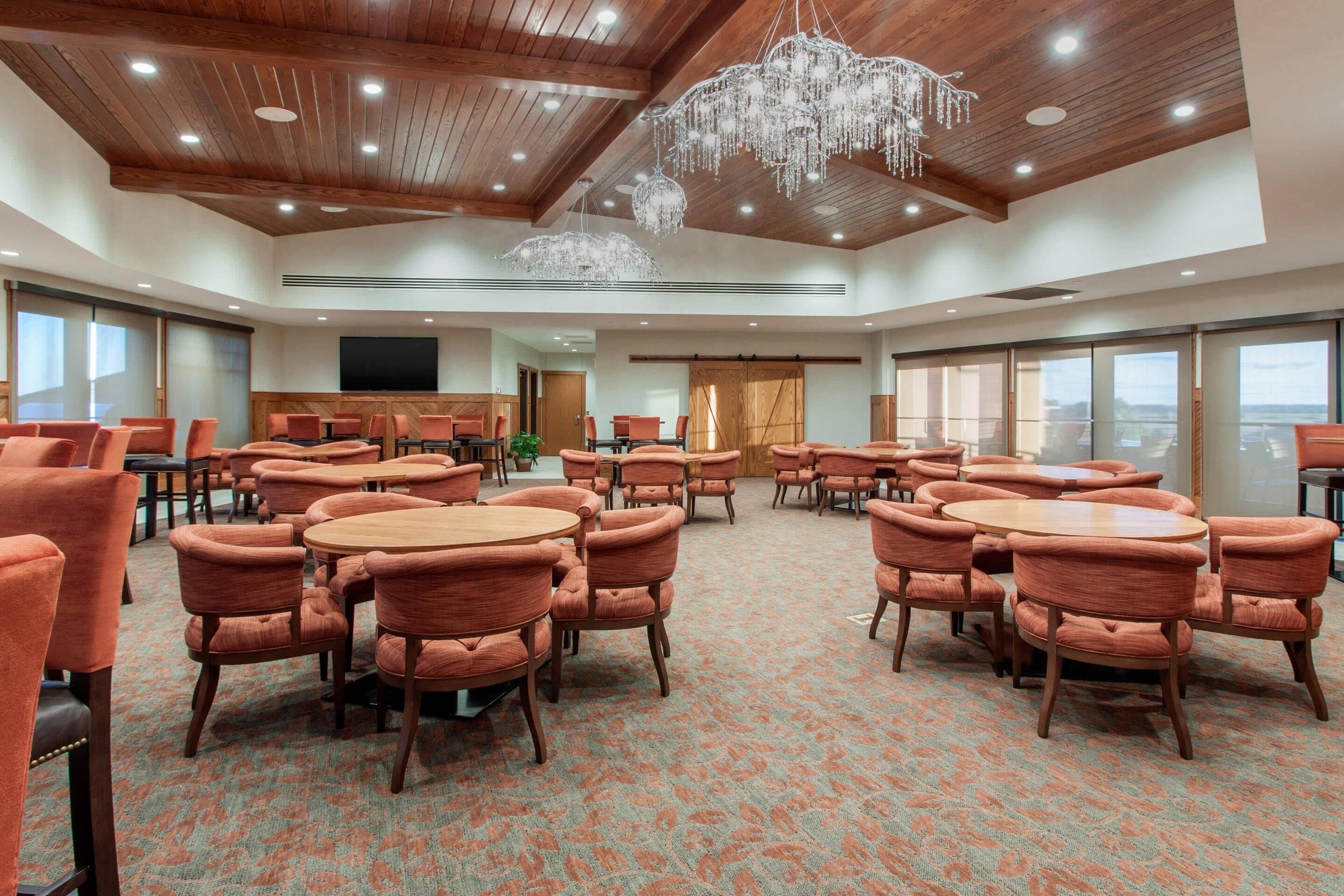 Une salle de conférence spacieuse, dotée de plusieurs tables rondes et de chaises recouvertes d’un tissu orange, de poutres en bois au plafond et d’un grand lustre.