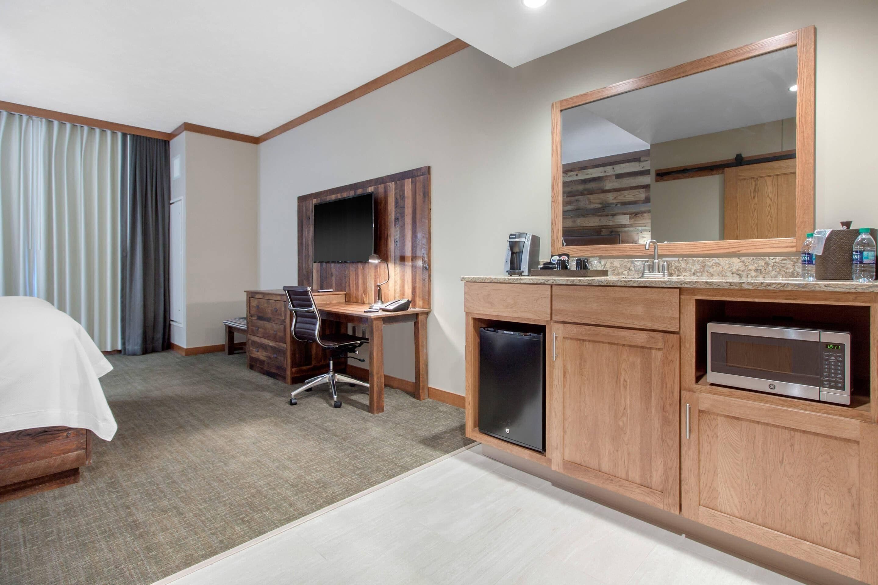 A hotel room with a bed, desk, chair, TV, microwave, and a large mirror.