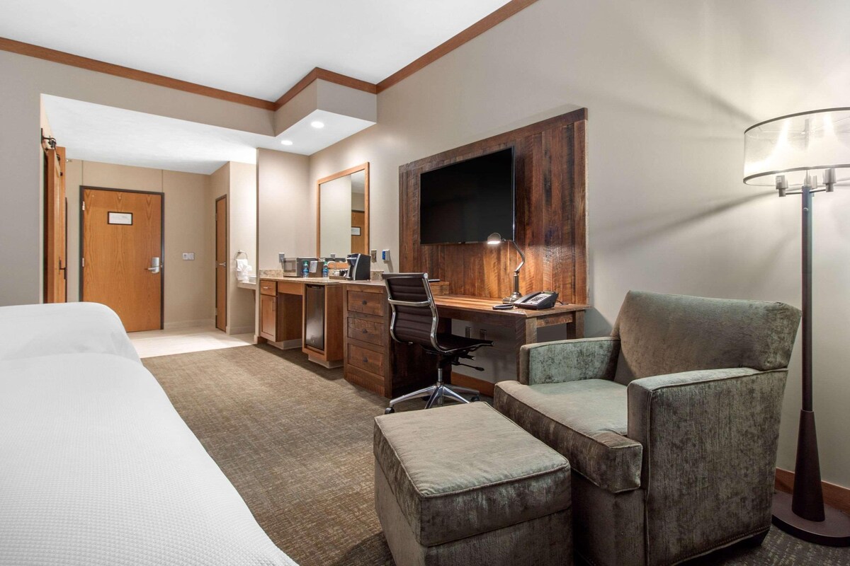 A hotel room with a bed, a desk with a chair, a TV mounted on the wall, and a floor lamp.