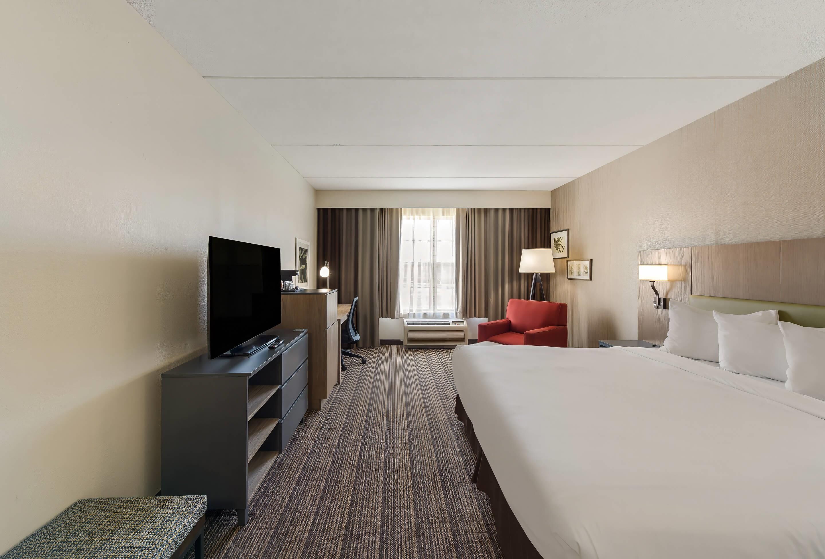 Ein Hotelzimmer mit einem großen Bett, einem Fernseher, einem Schreibtisch, einem Sessel und einem Fenster mit Vorhängen.
