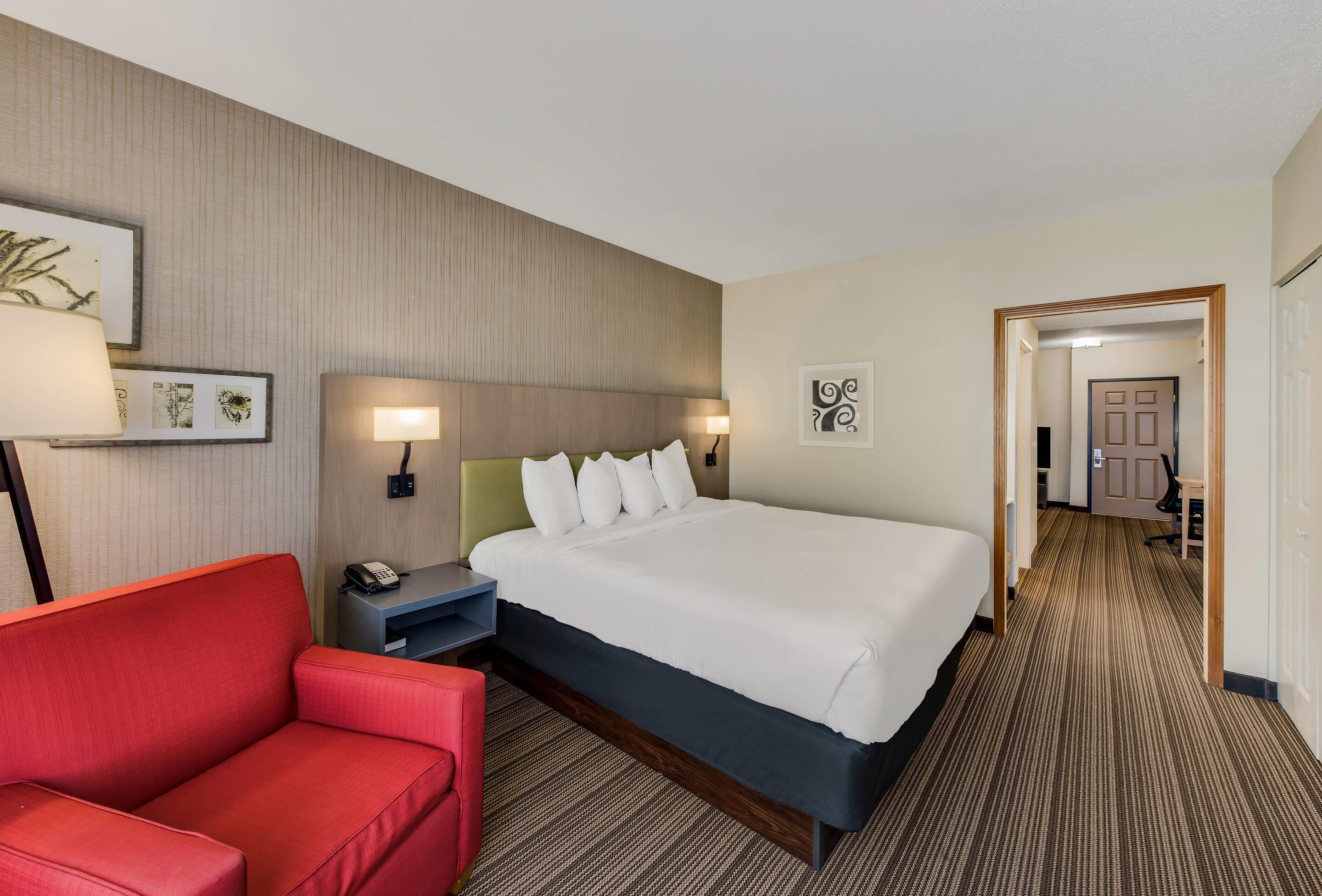 A hotel room with a large bed, a red armchair, a desk with a telephone, and a wall-mounted lamp.