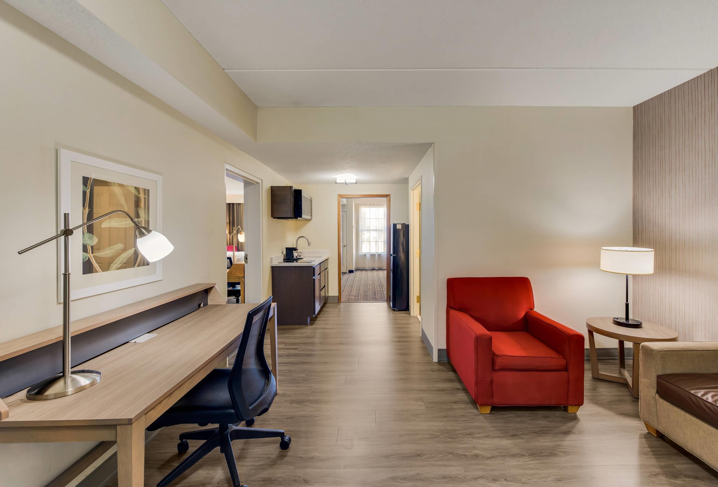 A hotel room with a wooden desk, a red armchair, a sofa, a TV mounted on the wall, and a small table with a lamp.
