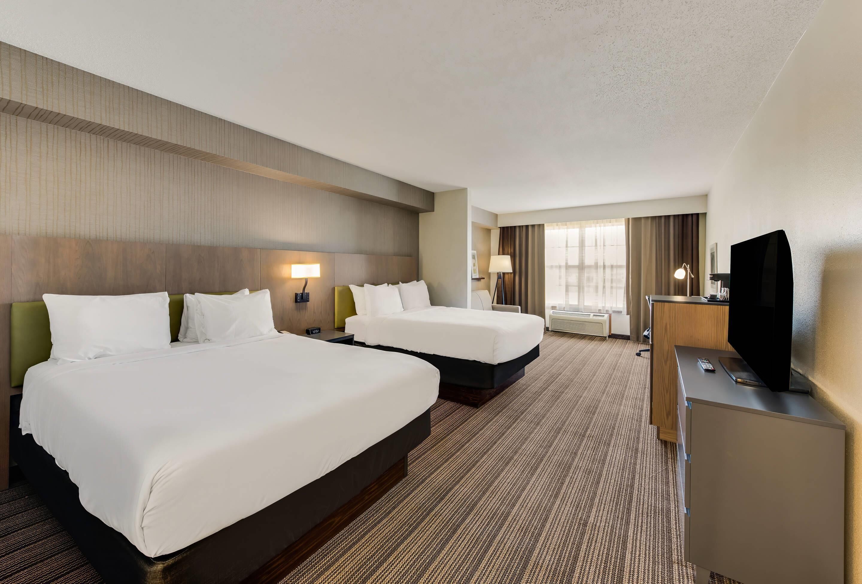 A hotel room with two beds, a desk, a television, and a large window with curtains.