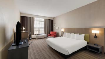 Une chambre d’hôtel avec un grand lit, un bureau avec une télévision, un fauteuil rouge, une table de chevet avec une lampe et une fenêtre avec des rideaux.