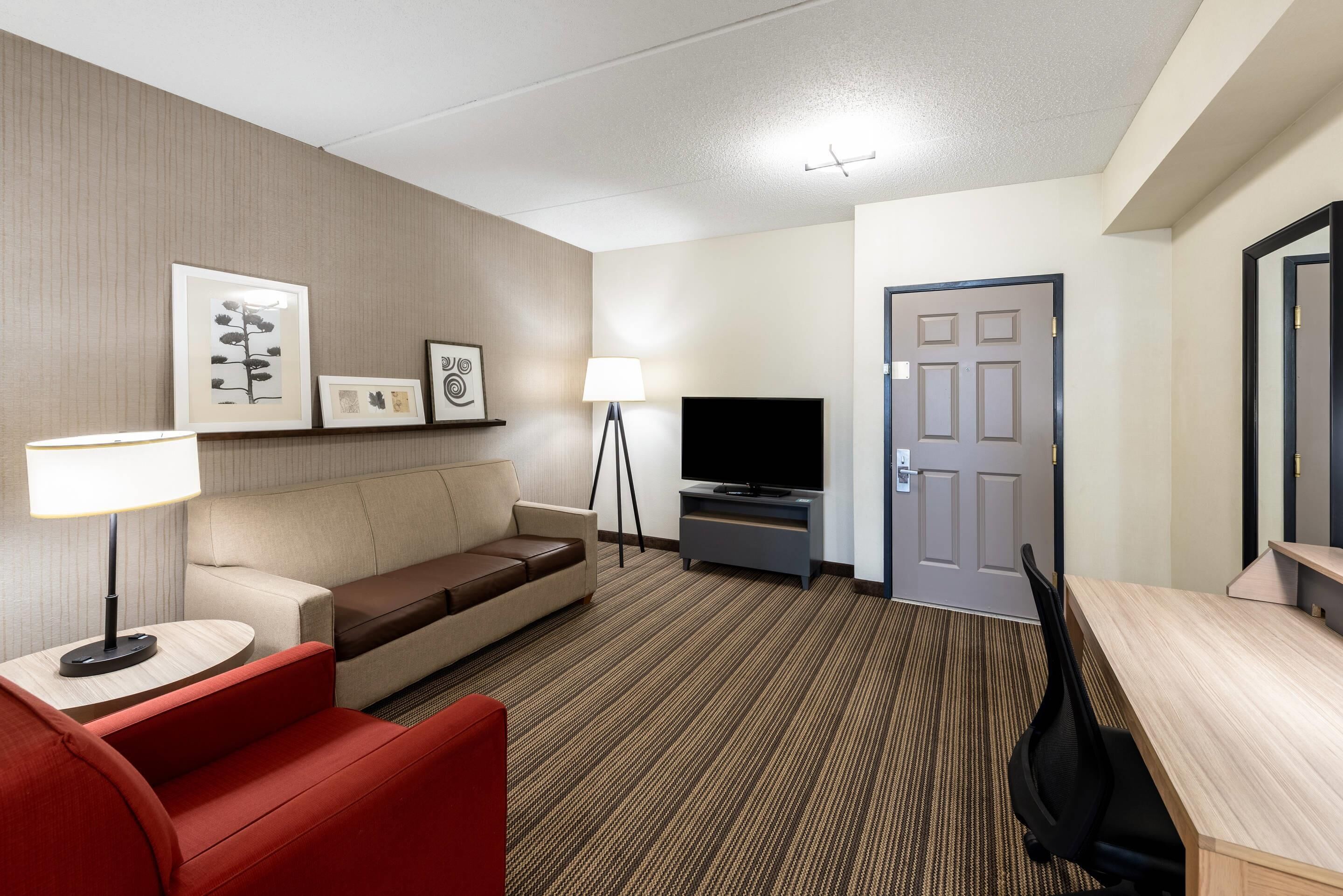 A hotel room with a sofa, a red armchair, a TV on a stand, a desk, and a door.