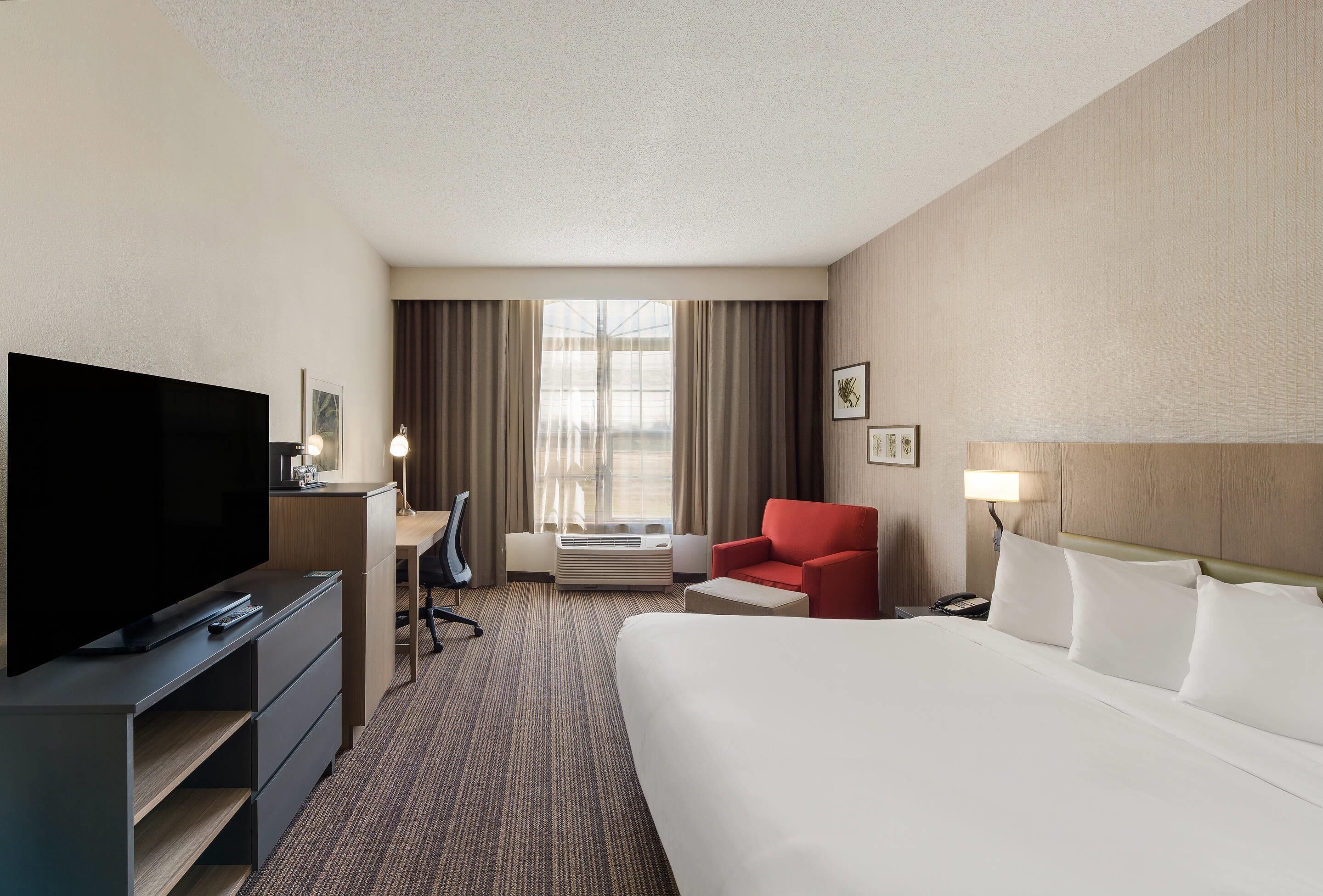 Una habitación de hotel con una cama grande, televisión, escritorio con silla, sillón rojo y ventana con cortinas.