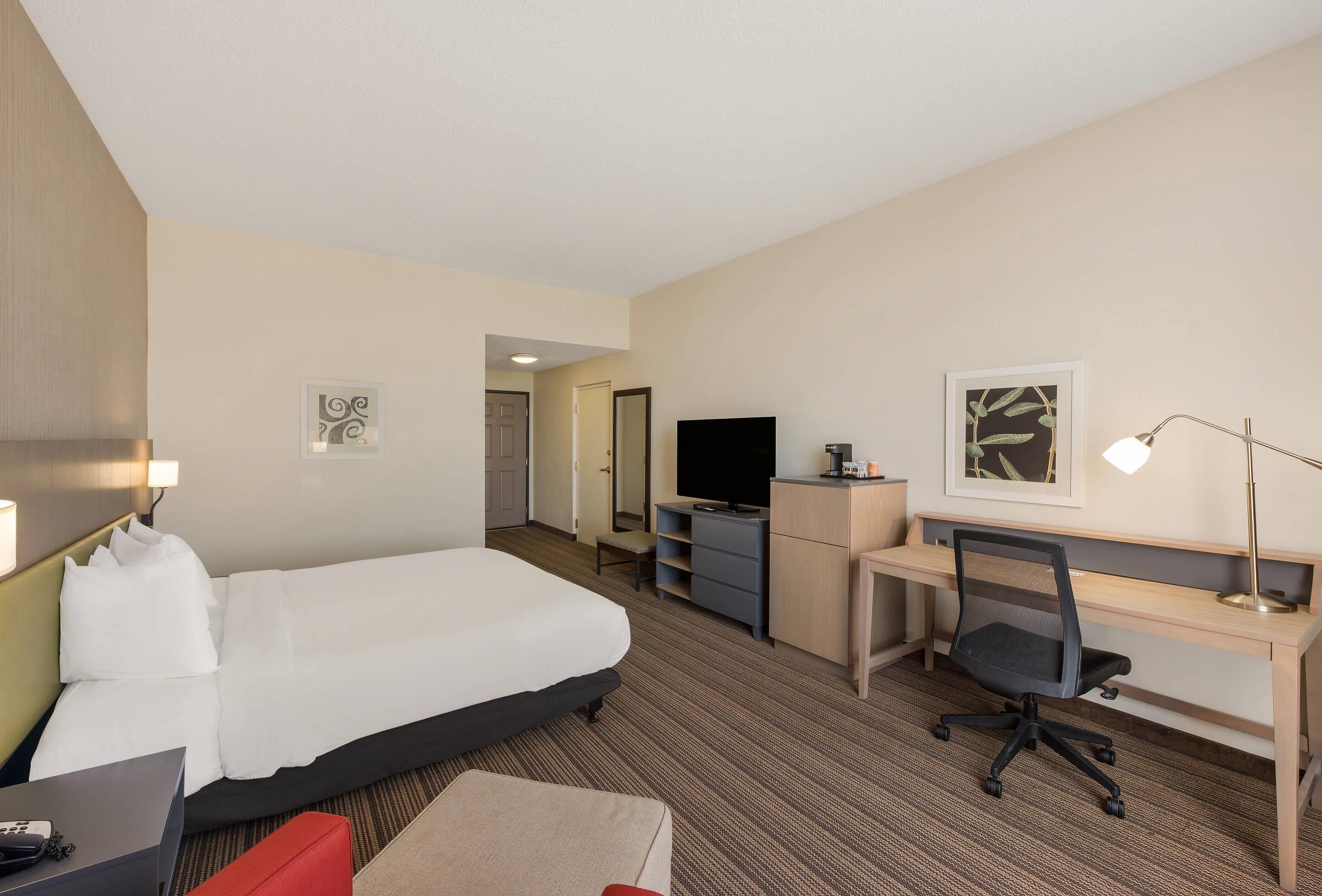 A hotel room with a bed, desk, chair, TV, and a small seating area.