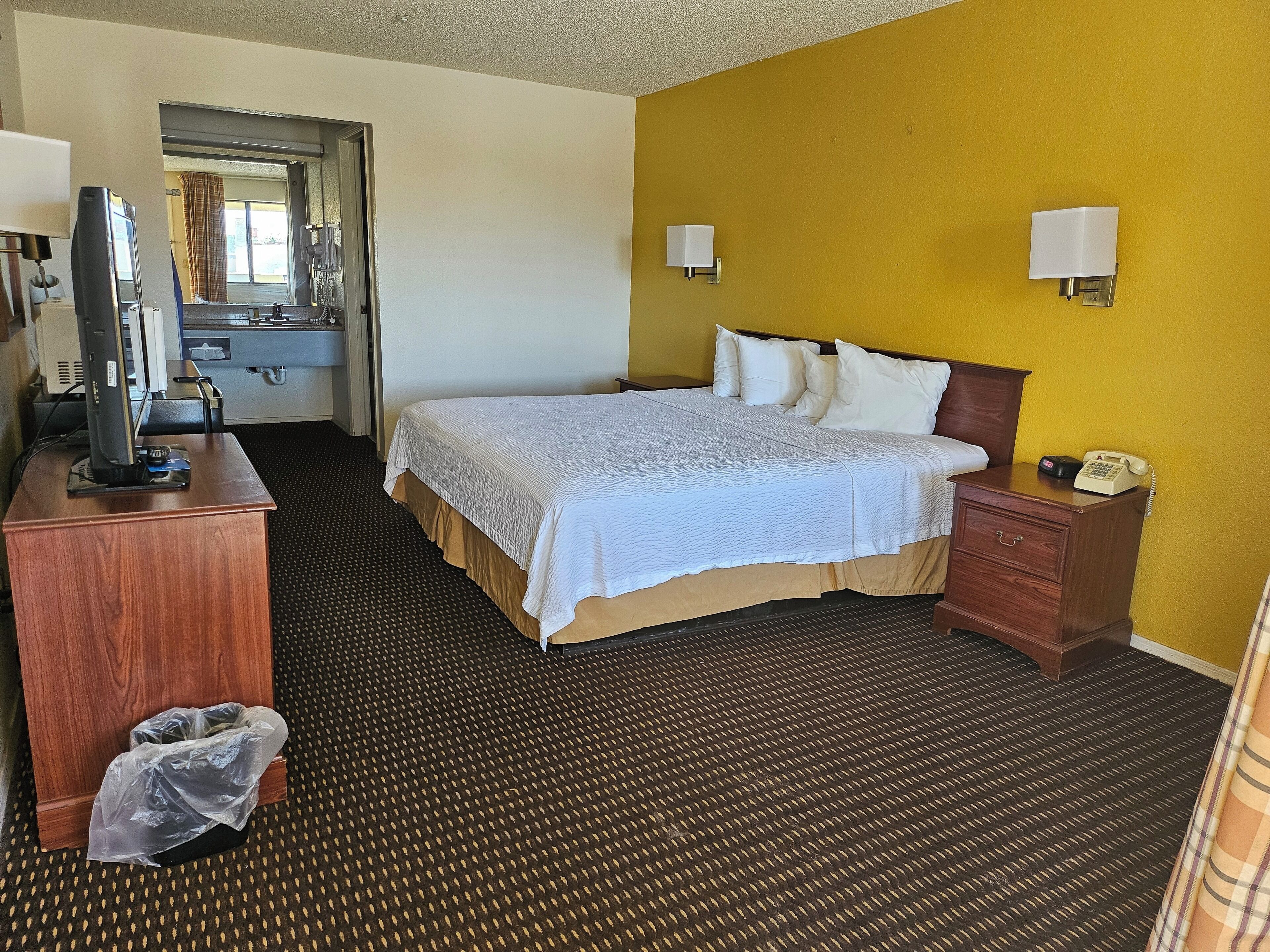 A hotel room with a bed, a desk with a telephone, a television, and a trash bin.