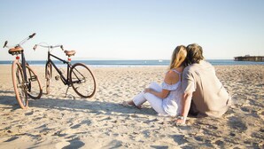 A couple sitting on a sandy beach with bicycles leaning against a wooden structure. - Avila Village Inn (Avila Beach)