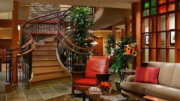 A spacious lobby with a curved staircase, a leather armchair, a sofa, and a coffee table with books and a vase of flowers. - Avila Village Inn (Avila Beach)