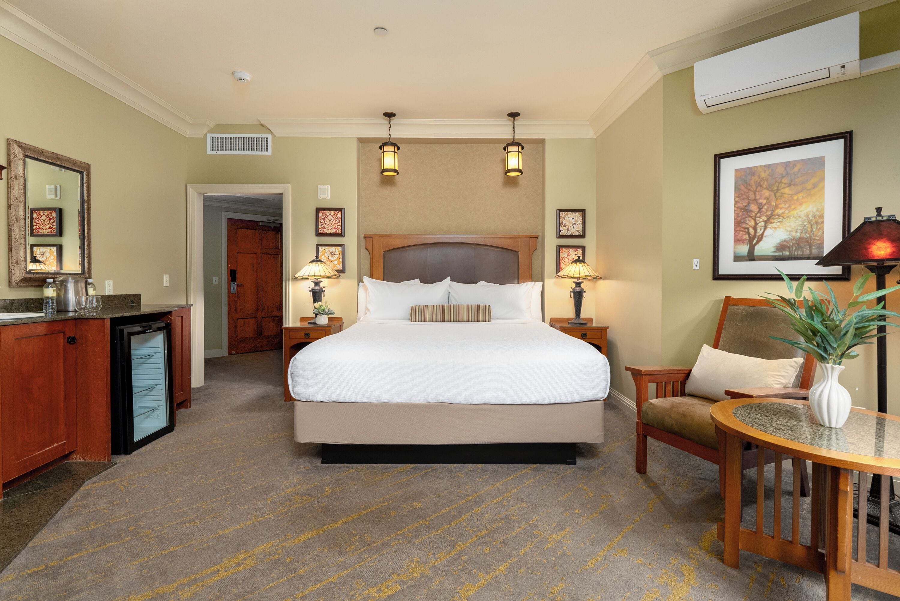 A hotel room with a large bed, bedside lamps, a desk with a lamp, a chair, and a small table with a plant.
