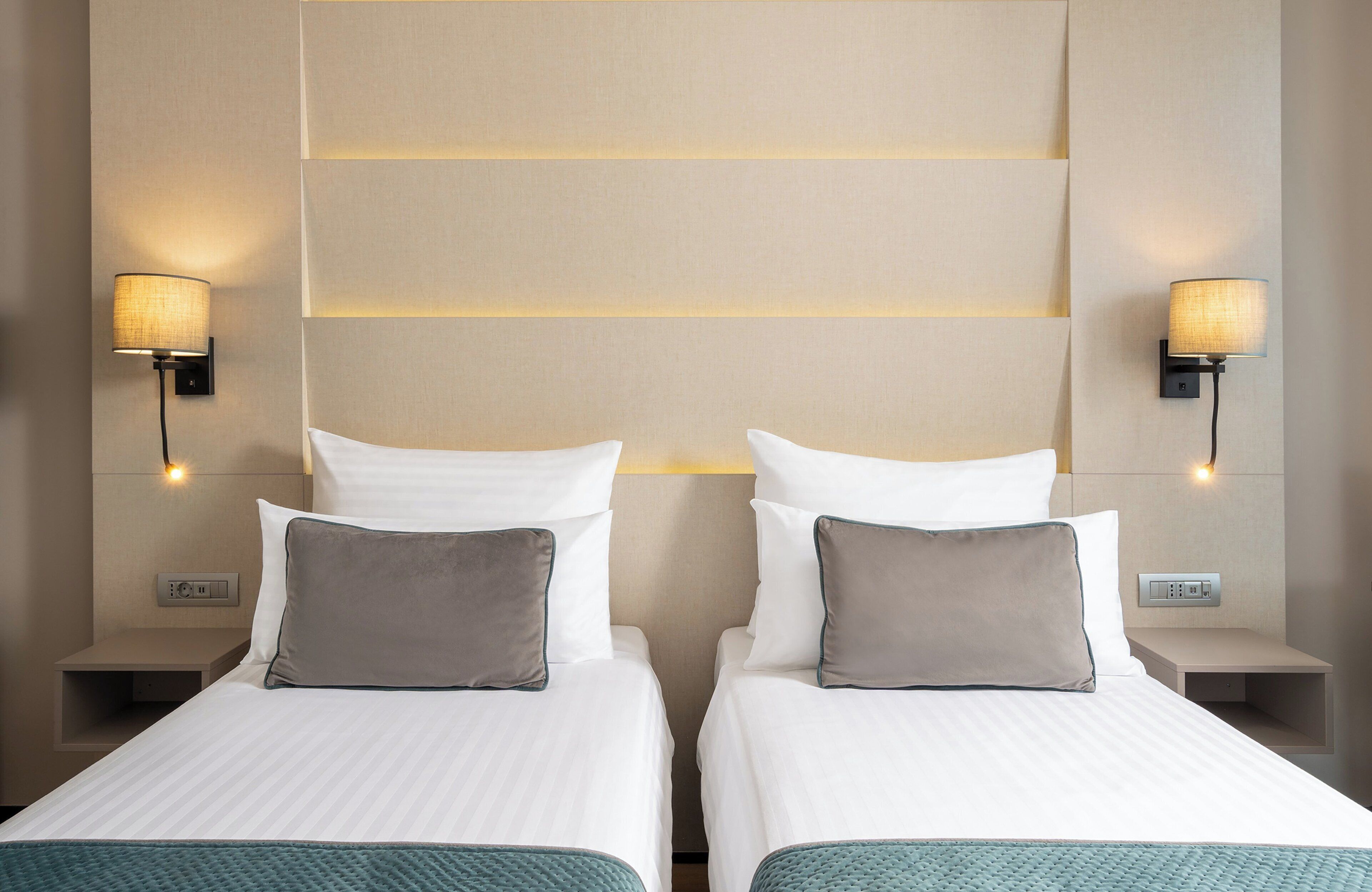 A hotel room with two beds, each with white linens and a teal throw. There are wall-mounted lamps on either side of the beds, and a control panel on the wall between them.