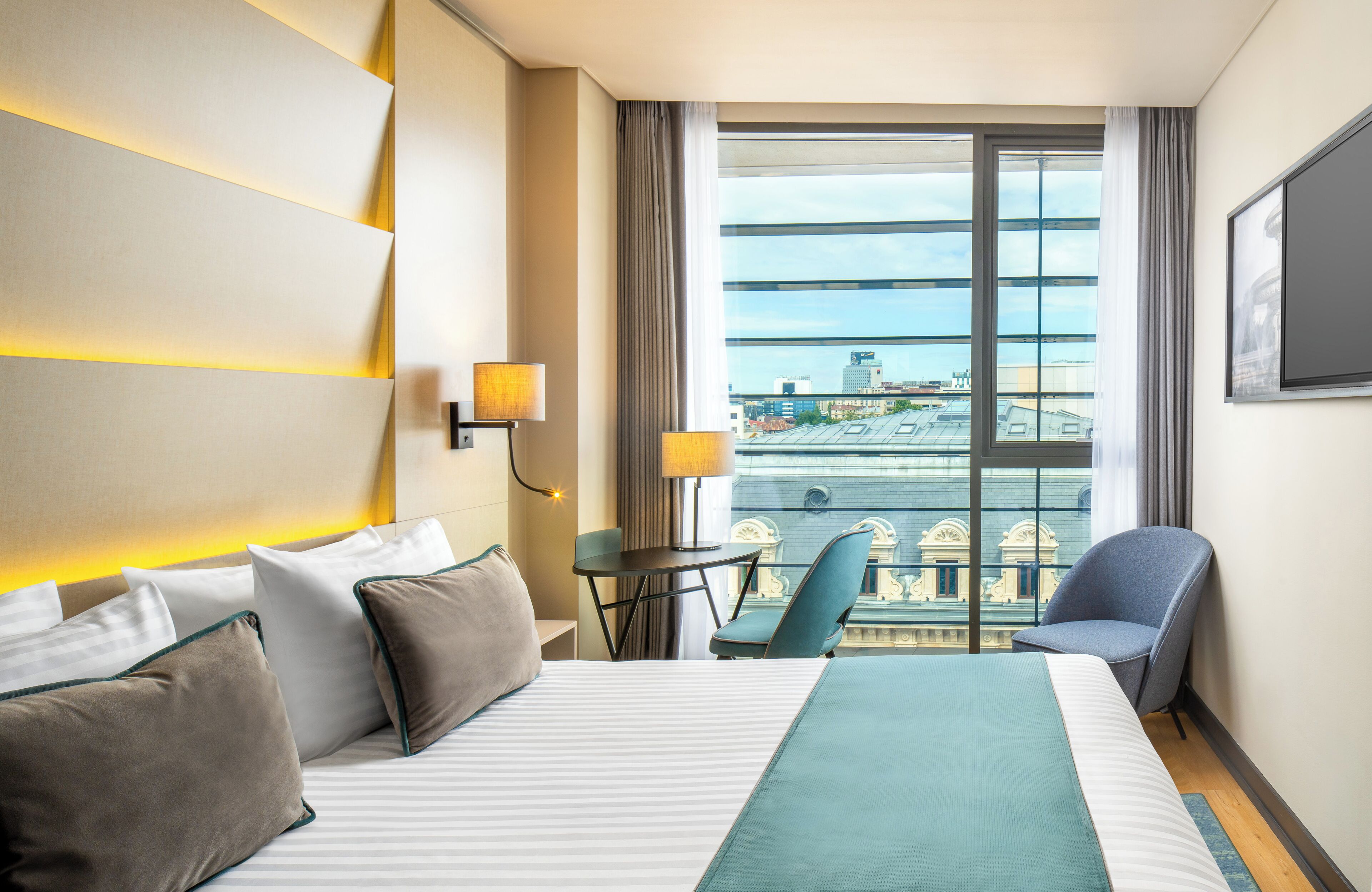 Une chambre d’hôtel avec un grand lit, deux fauteuils, une petite table et une vue sur la ville grâce à une grande fenêtre.