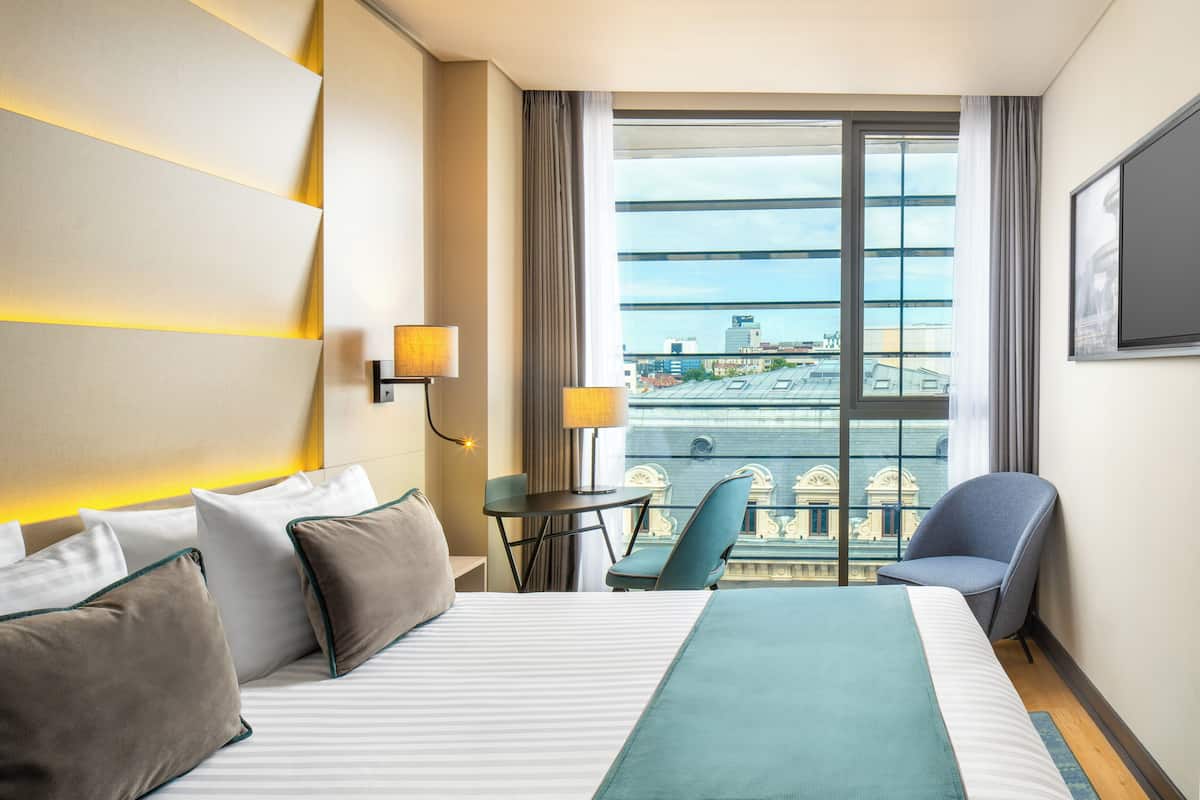 A hotel room with a large bed, two chairs, a small table, and a view of the city through a large window.