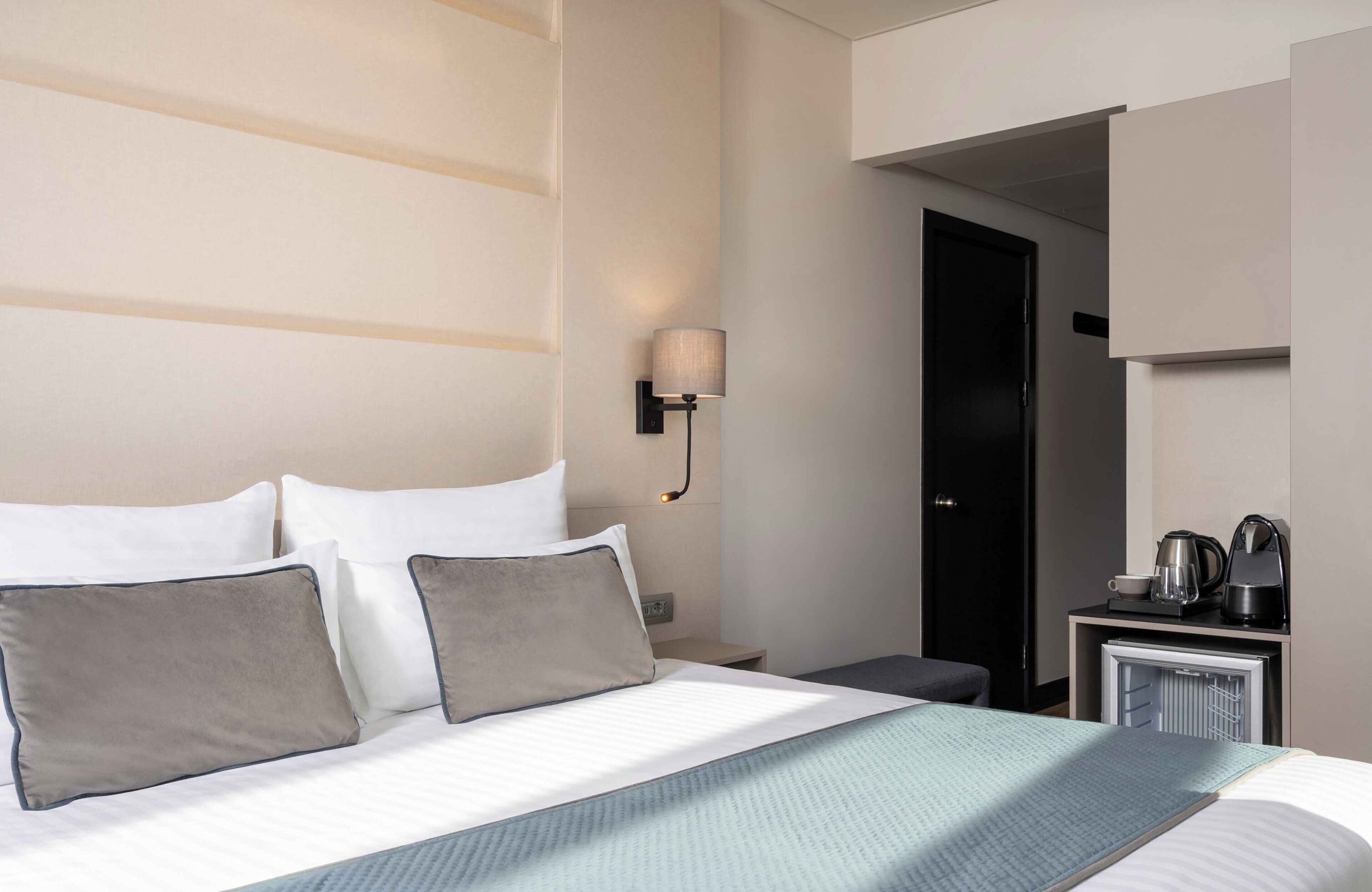 A hotel room with a bed, a nightstand, a lamp, a coffee maker, and a mini-fridge.