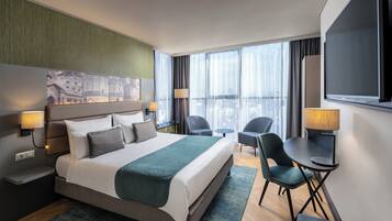 Una habitación de hotel moderna con una cama grande, dos mesitas de noche, un escritorio con silla, un televisor y un ventanal con cortinas.