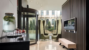 Una habitación de hotel moderna con un amplio ventanal, una mesa de vidrio con fruta, dos sillas tapizadas de verde, un banco y un televisor de pantalla plana montado sobre un panel negro.