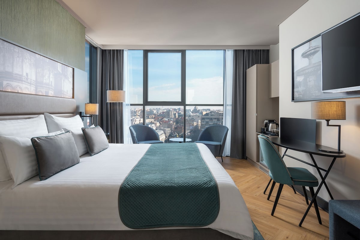 Habitación de hotel con cama, escritorio, dos sillas, televisor y ventana grande con vista a la ciudad.