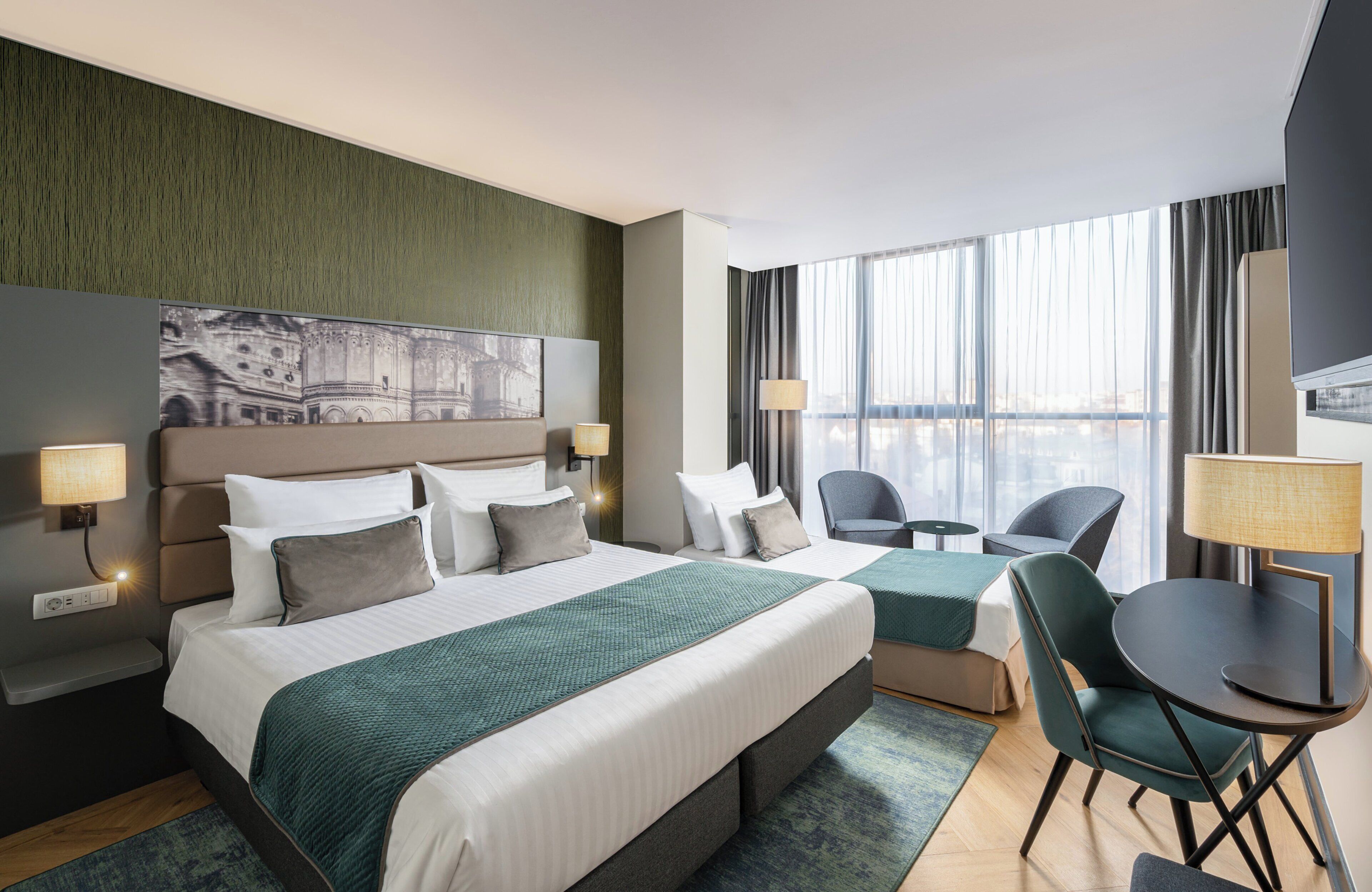 Una habitación de hotel moderna con una cama grande, dos mesitas de noche con lámparas, un espacio de descanso con una mesa y sillas, y un ventanal con cortinas.