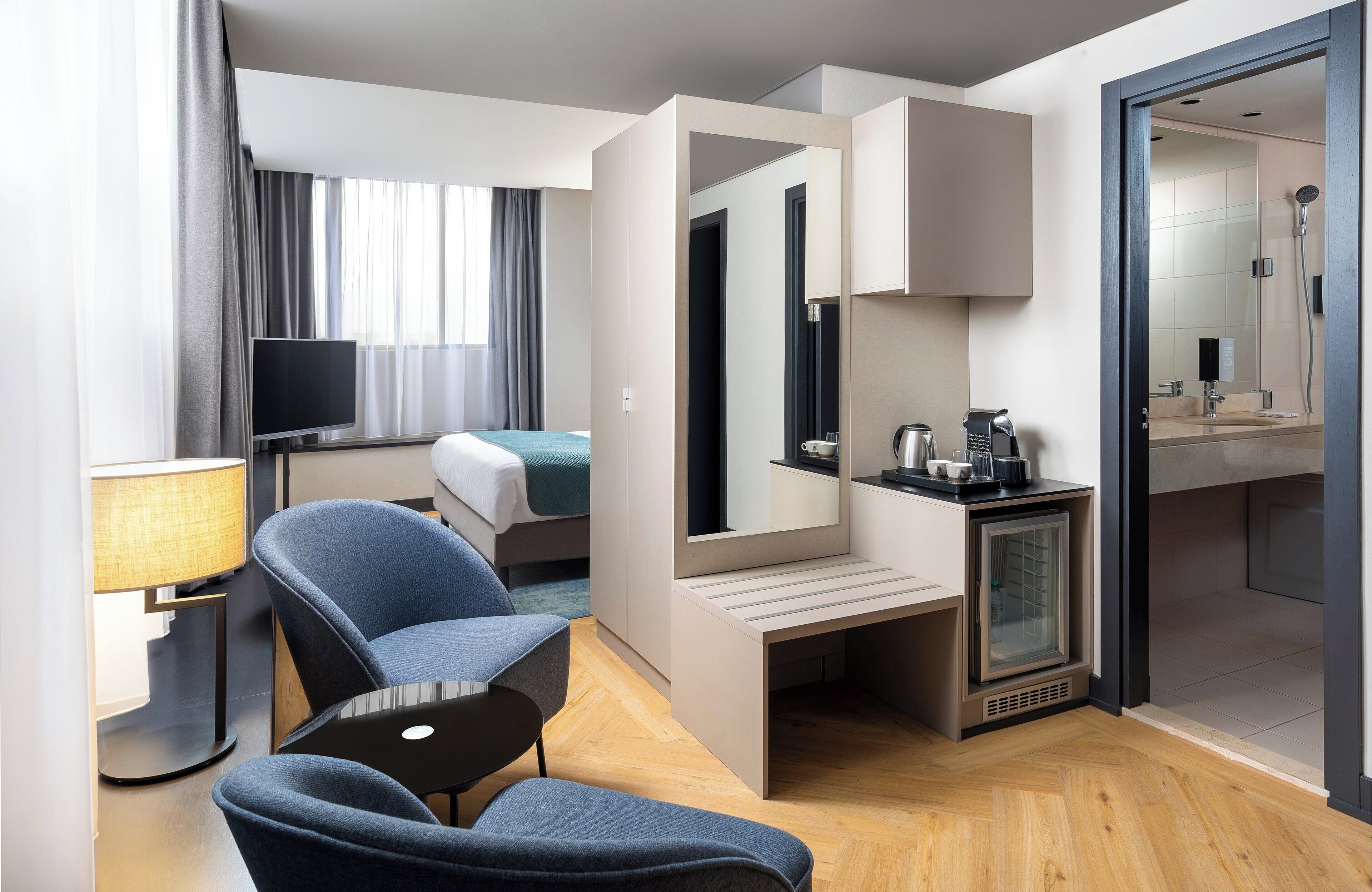 Een moderne hotelkamer met een bed, een zithoek met stoelen, een televisie, een spiegel, een minibar en een badkamer die door een geopende deur zichtbaar is.