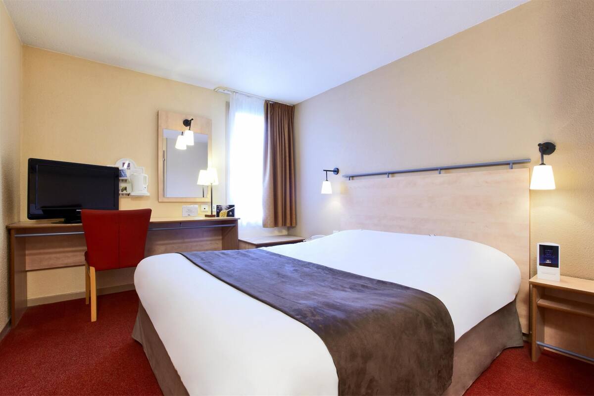Quarto de hotel com uma cama, uma escrivaninha com televisão, uma cadeira vermelha, um espelho e uma mesa de cabeceira com abajur.
