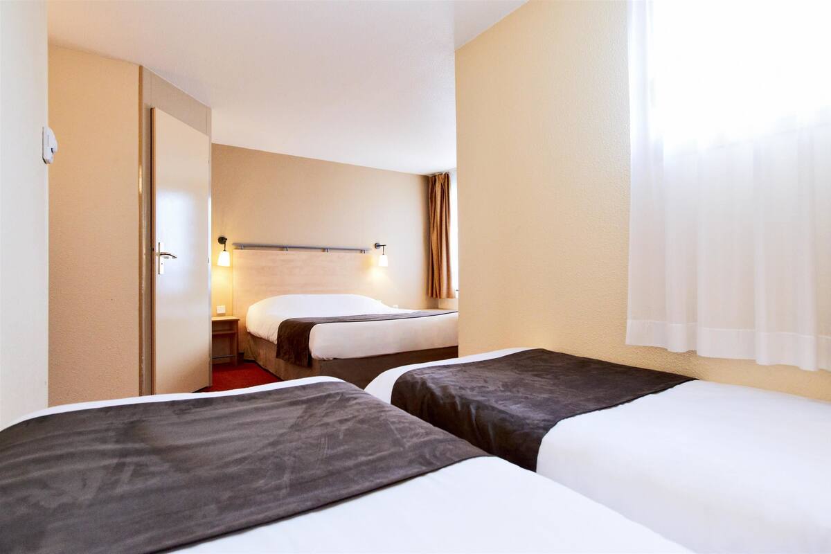 Quarto de hotel com duas camas, uma mesa de cabeceira, uma luminária e uma janela com cortinas.