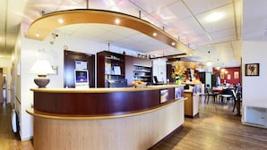 A reception area with a curved wooden counter, a lamp, a coffee machine, and a display of pastries. - Hotel Kyriad Beauvais Sud (Beauvais)