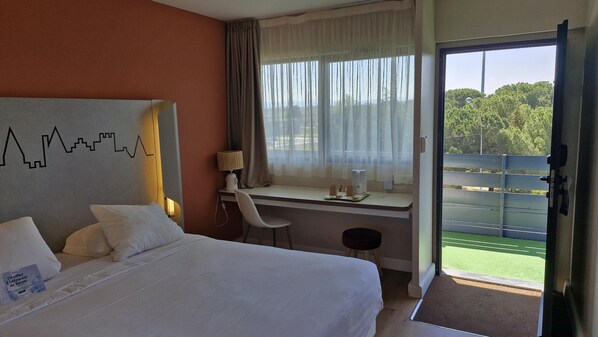 A hotel room with a bed, a desk with a chair, a lamp, and a view of a green landscape through a large window.