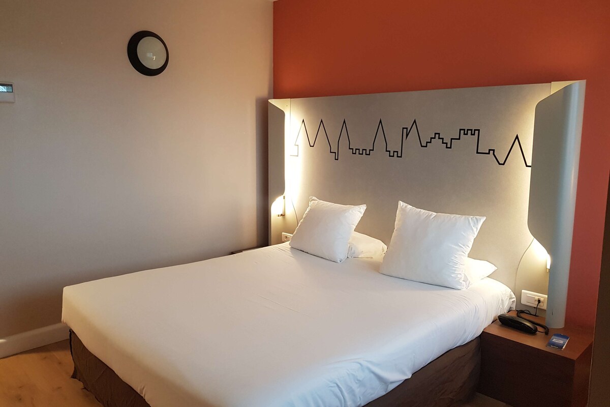 Una cama bien tendida con sábanas blancas y dos almohadas, una mesita de noche con un teléfono y un libro, y una lámpara de pared con diseño geométrico.