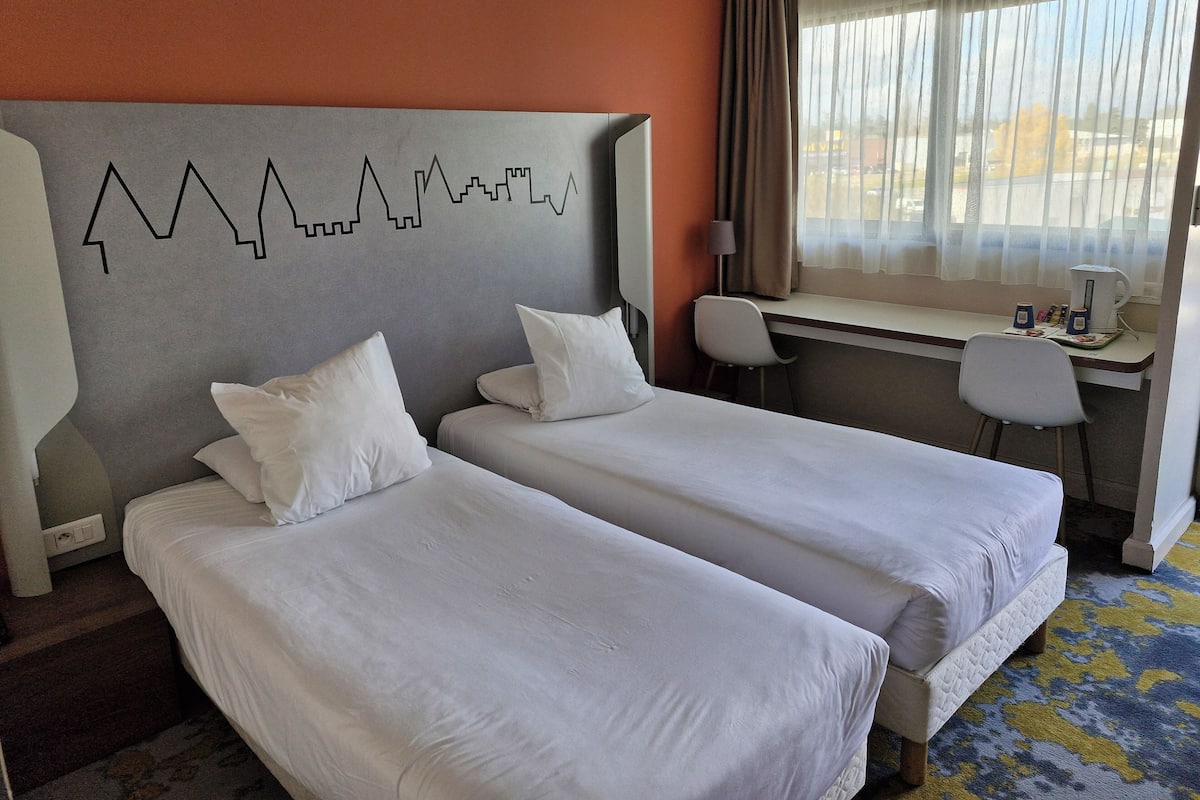 Habitación de hotel con dos camas, un escritorio, una silla y una ventana con cortinas.