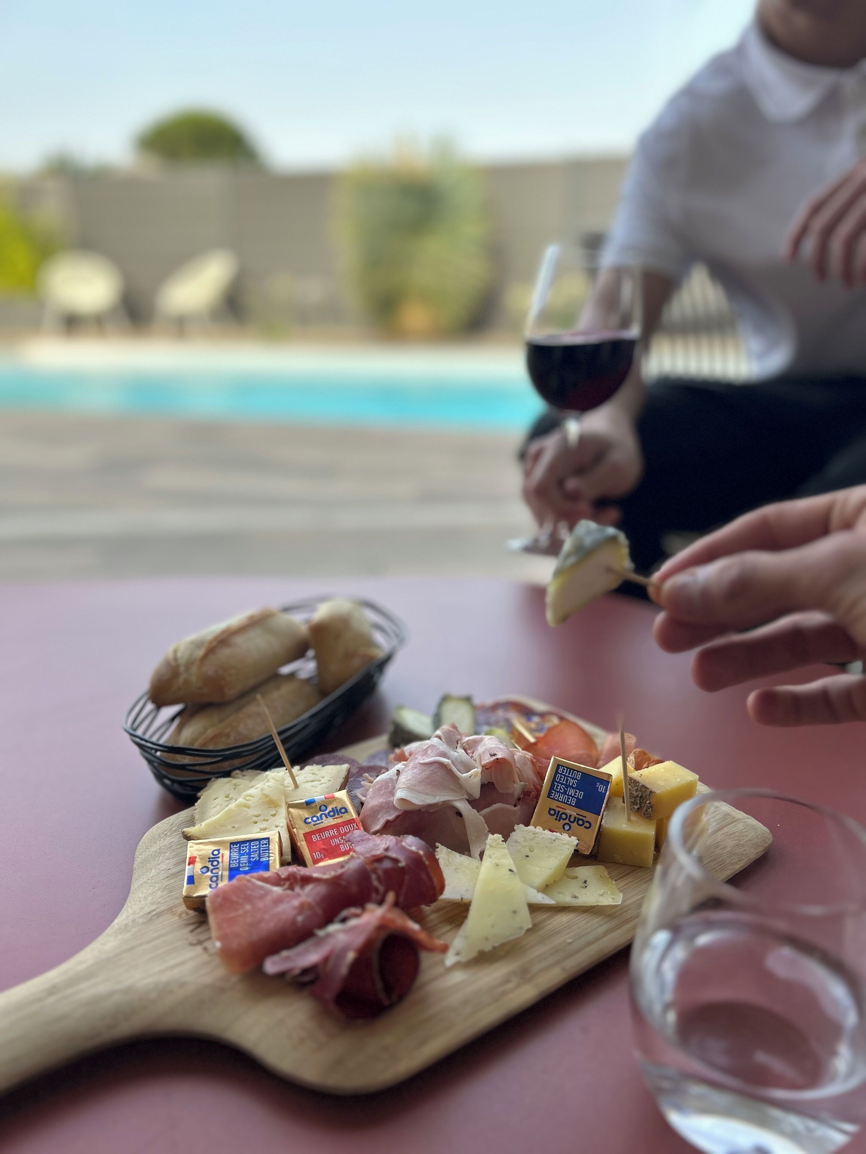 A wooden board with various cheeses and cured meats, a glass of red wine, and a person holding a piece of cheese.
