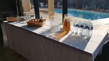Une table installée au bord d’une piscine, avec un panier de croissants, une carafe de thé glacé et plusieurs bouteilles d’eau.