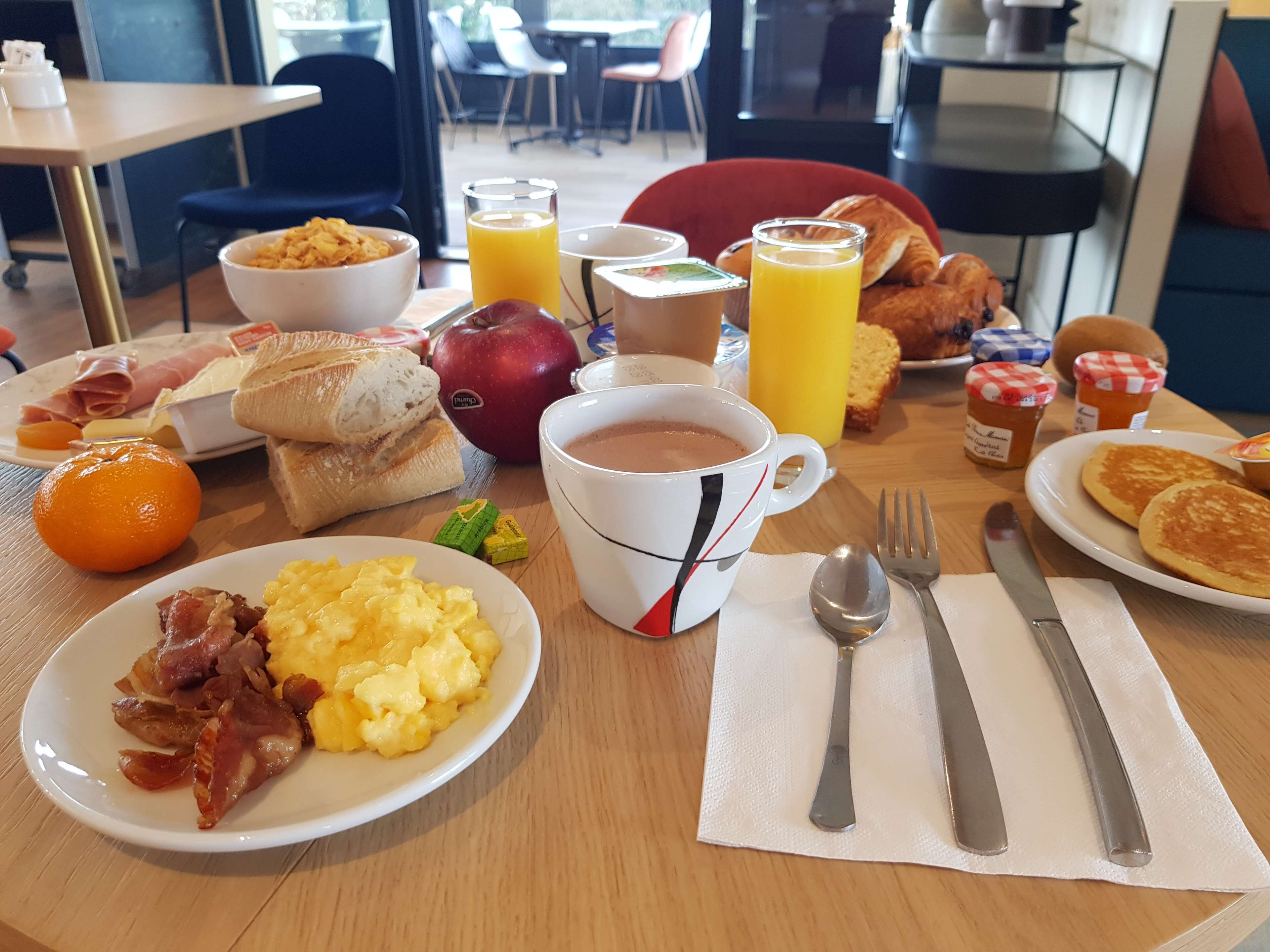A breakfast table with plates of eggs, bacon, toast, and pastries, a mug of coffee, and a glass of orange juice.