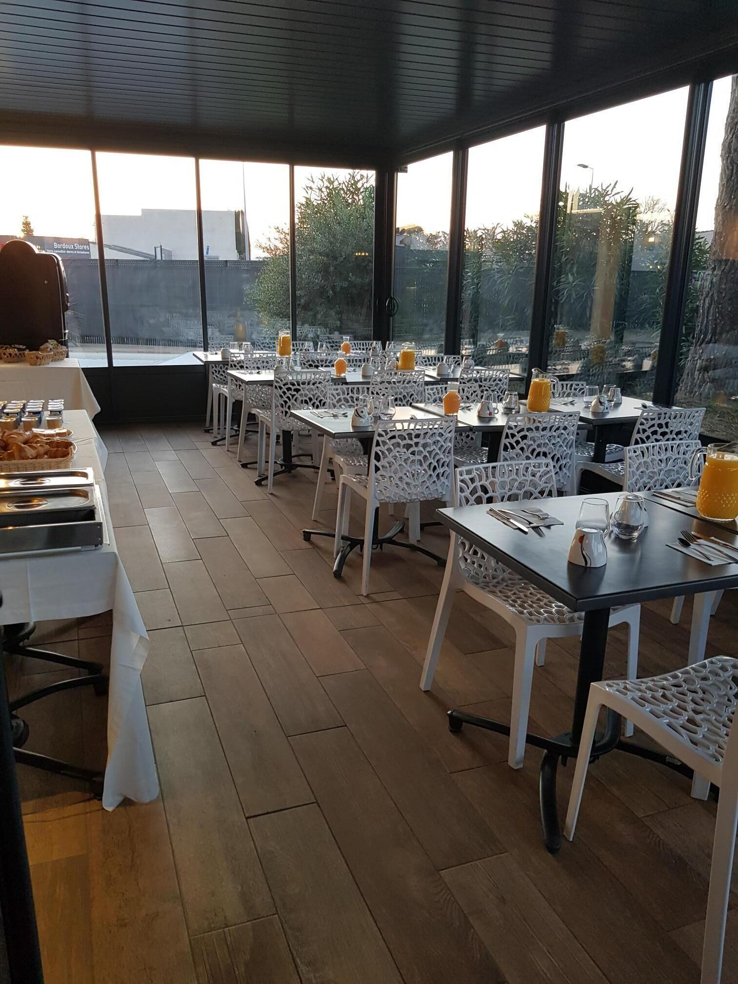 Een terras met witte stoelen en tafels, een buffet met eten, en uitzicht op bomen en gebouwen door grote ramen.