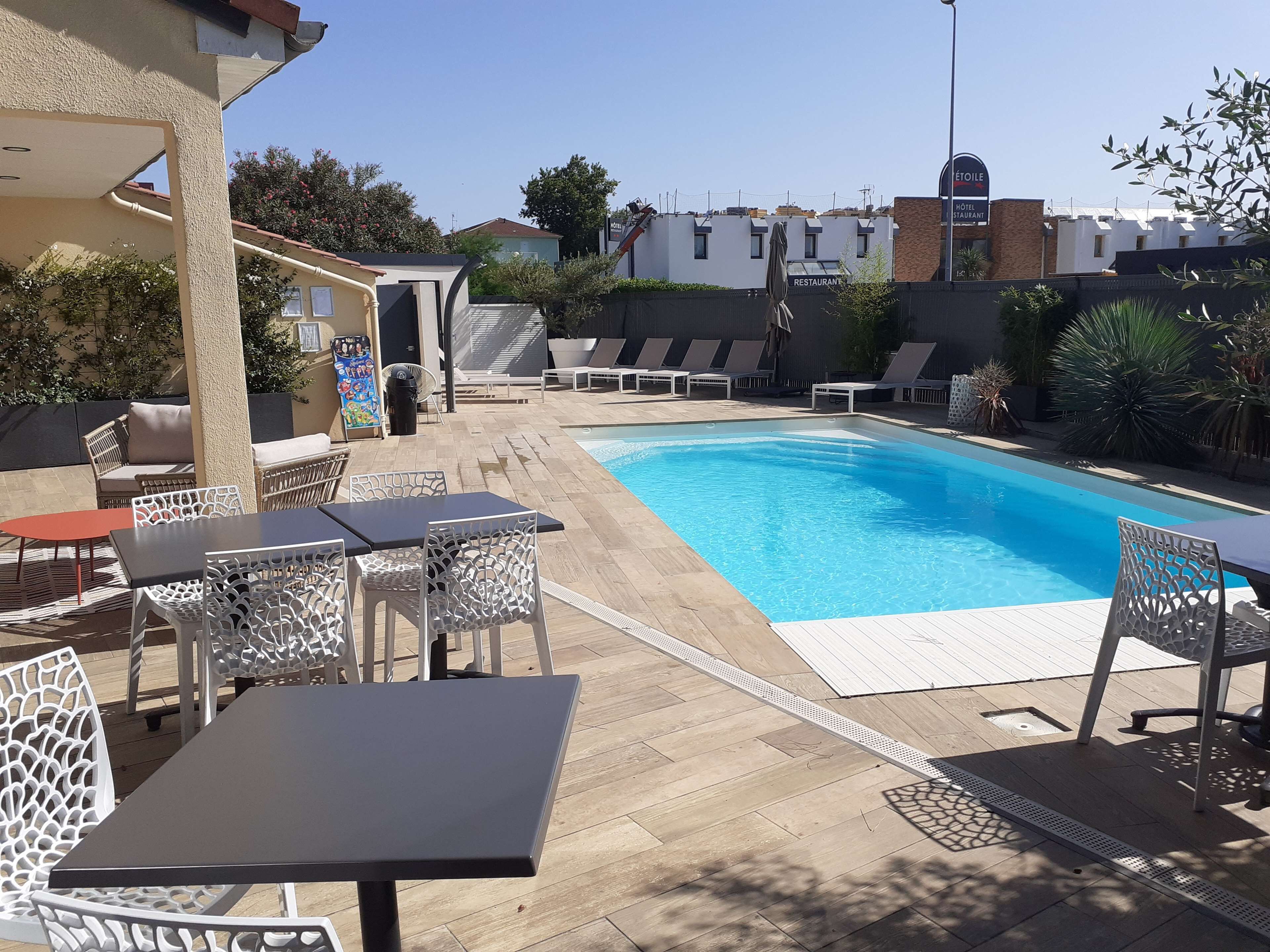 A pool area with lounge chairs and tables, surrounded by a low wall and vegetation.
