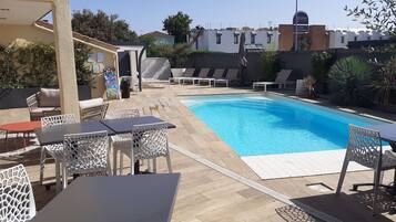A pool area with lounge chairs and tables, surrounded by a low wall and vegetation.