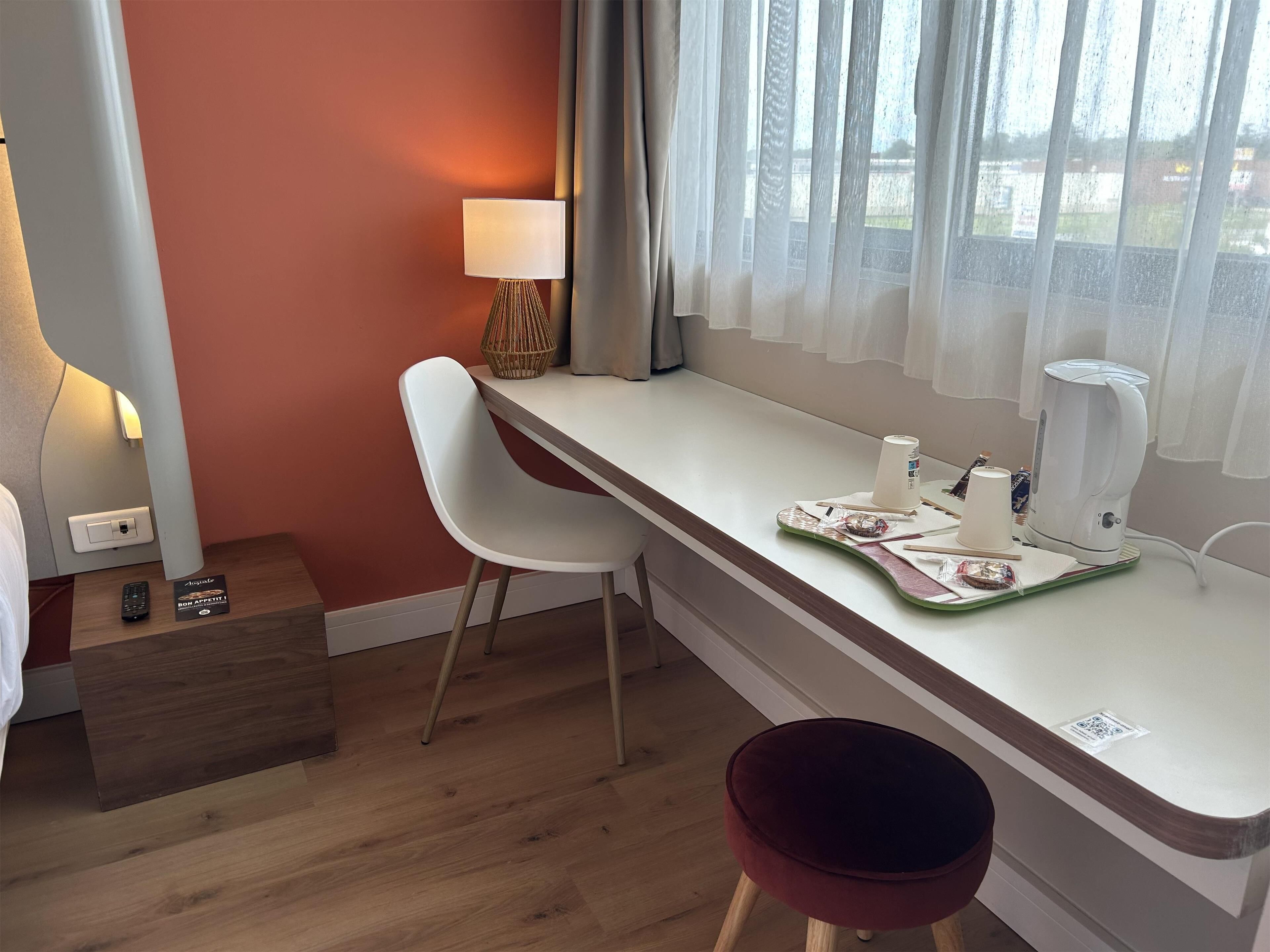 A modern hotel room with a wooden floor, a white desk with a lamp, a red chair, and a view of the outdoors through sheer curtains.