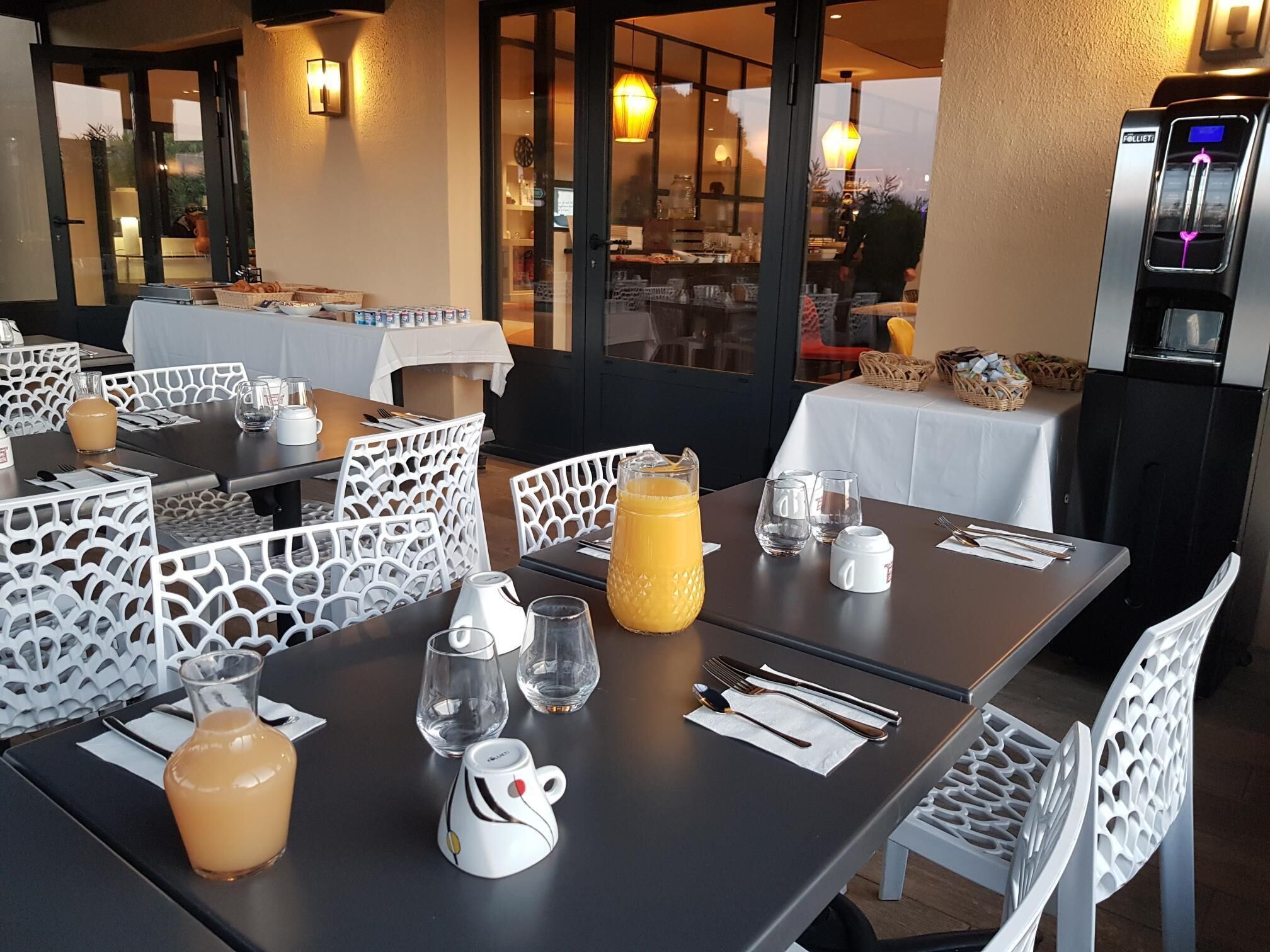 Un área de comedor al aire libre con mesas dispuestas para dos, que cuenta con sillas blancas con detalles recortados. Cada mesa tiene un mantel blanco, una jarra de jugo de naranja y una taza blanca.