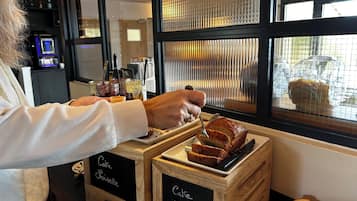 A person serving cake from a wooden crate labeled "Café Sloisette" and "Café Vanille" in a café setting with a window view.