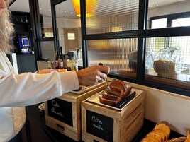 A person serving cake from a wooden crate labeled "Café Sloisette" and "Café Vanille" in a café setting with a window view.