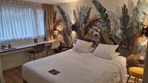 Ett hotellrum med en säng, ett skrivbord, en stol, en lampa och en väggmålning med tropiska blad.