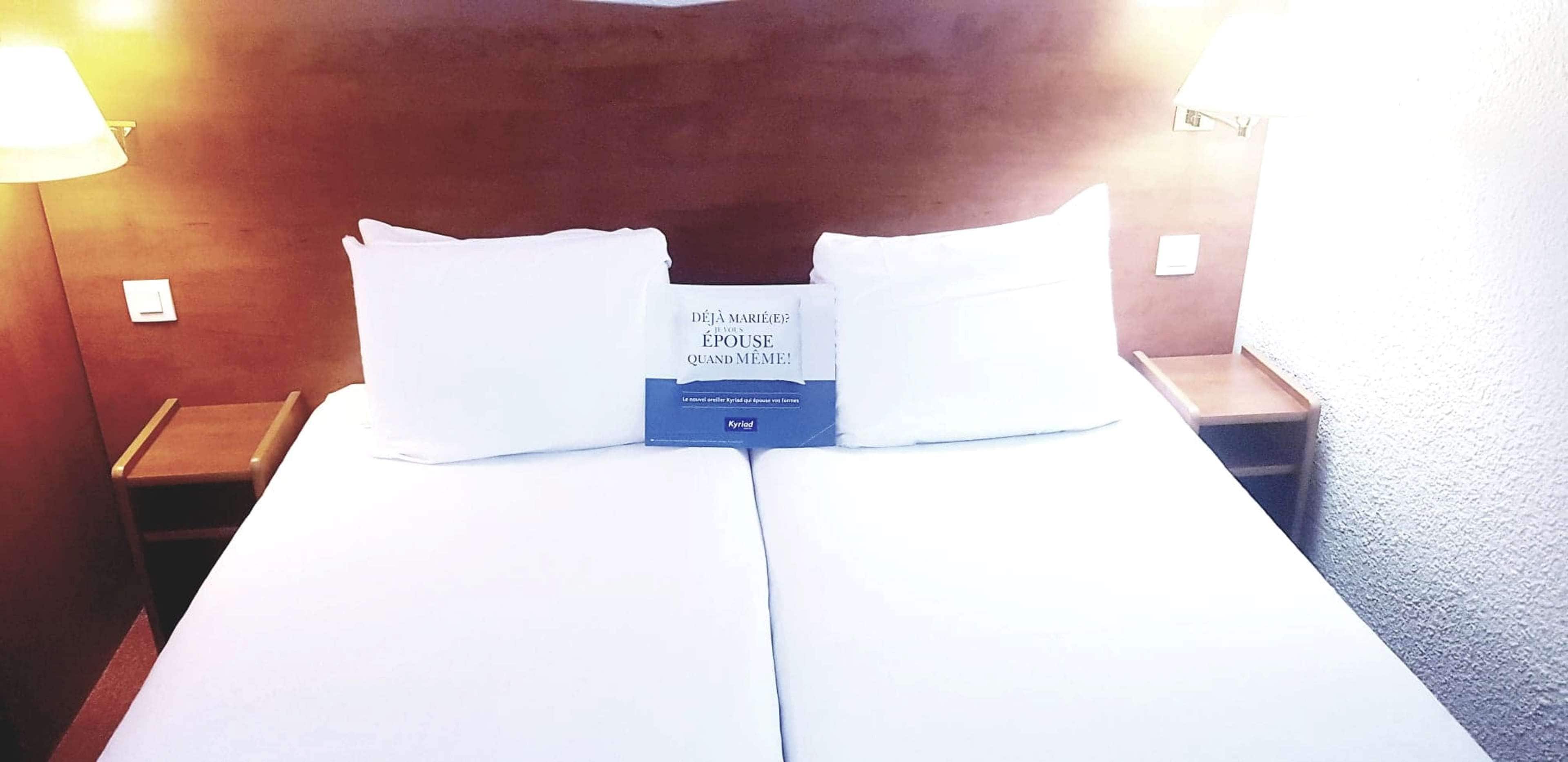 A hotel room with a bed, two pillows, a nightstand, and a wall-mounted lamp.