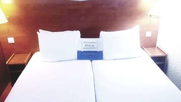 A hotel room with a bed, two pillows, a nightstand, and a wall-mounted lamp.