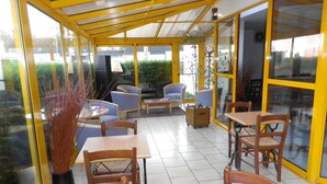 A covered outdoor seating area with tables and chairs, large windows, and a view of the outdoors. - Kyriad Chantilly (Gouvieux)
