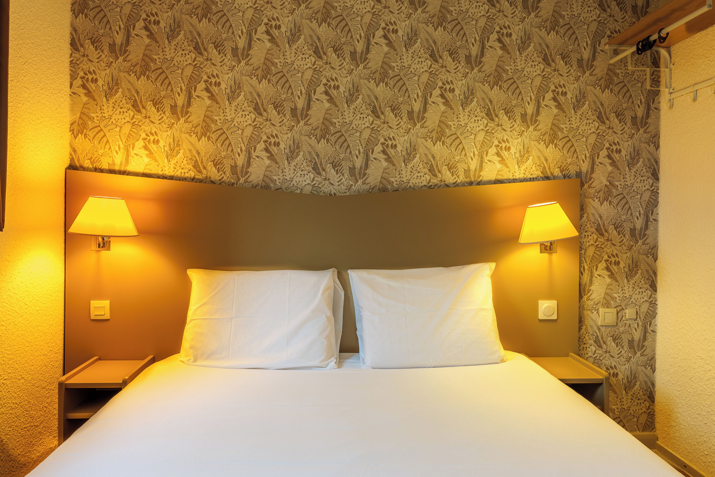 A bed with two pillows, a nightstand, a lamp, and a wall with a floral pattern.