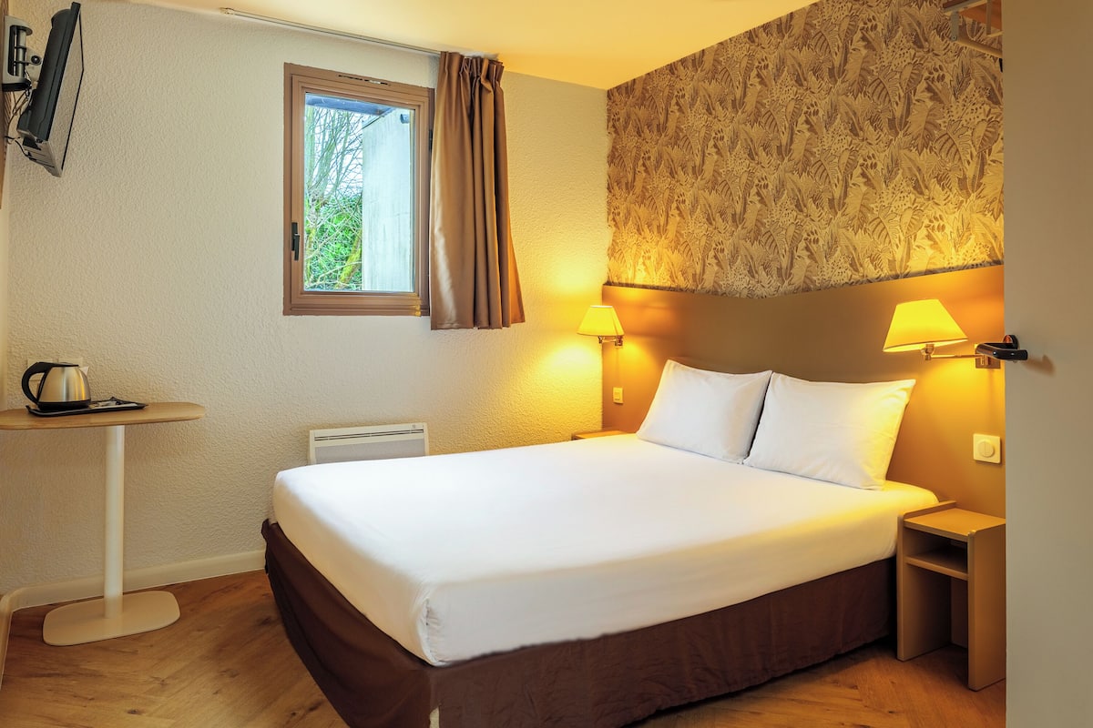 Une chambre d’hôtel avec un lit, deux lampes de chevet, une petite table avec une bouilloire, une fenêtre avec des rideaux et un mur décoré d’un motif.