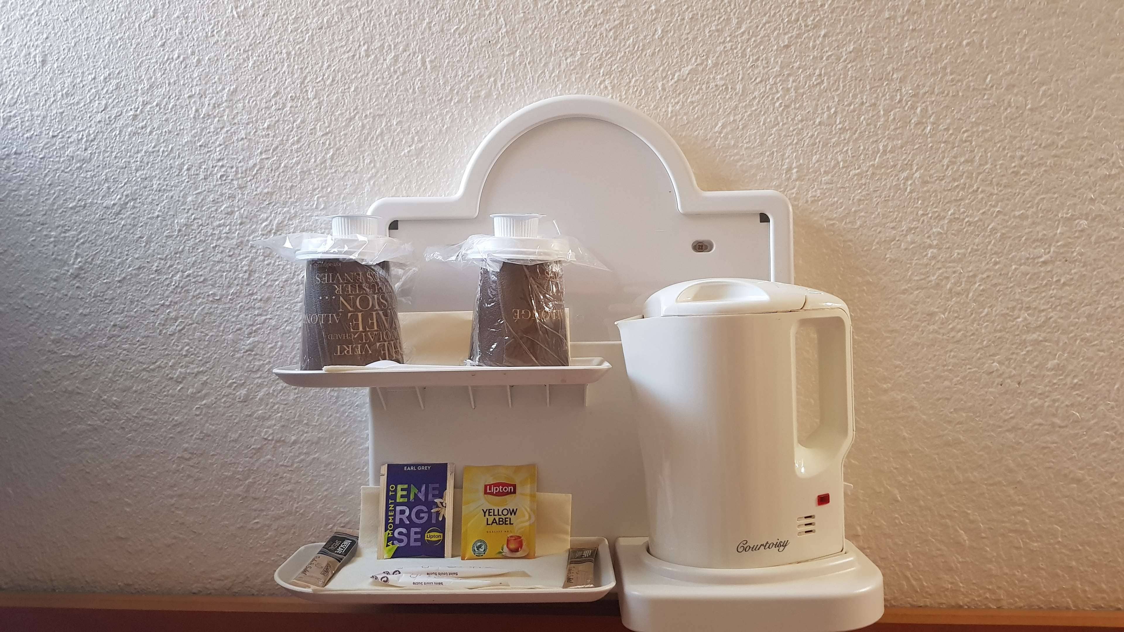 Un coin cuisine avec un plan de travail blanc, une machine à café et une étagère contenant des bocaux et une boîte de thé Lipton.